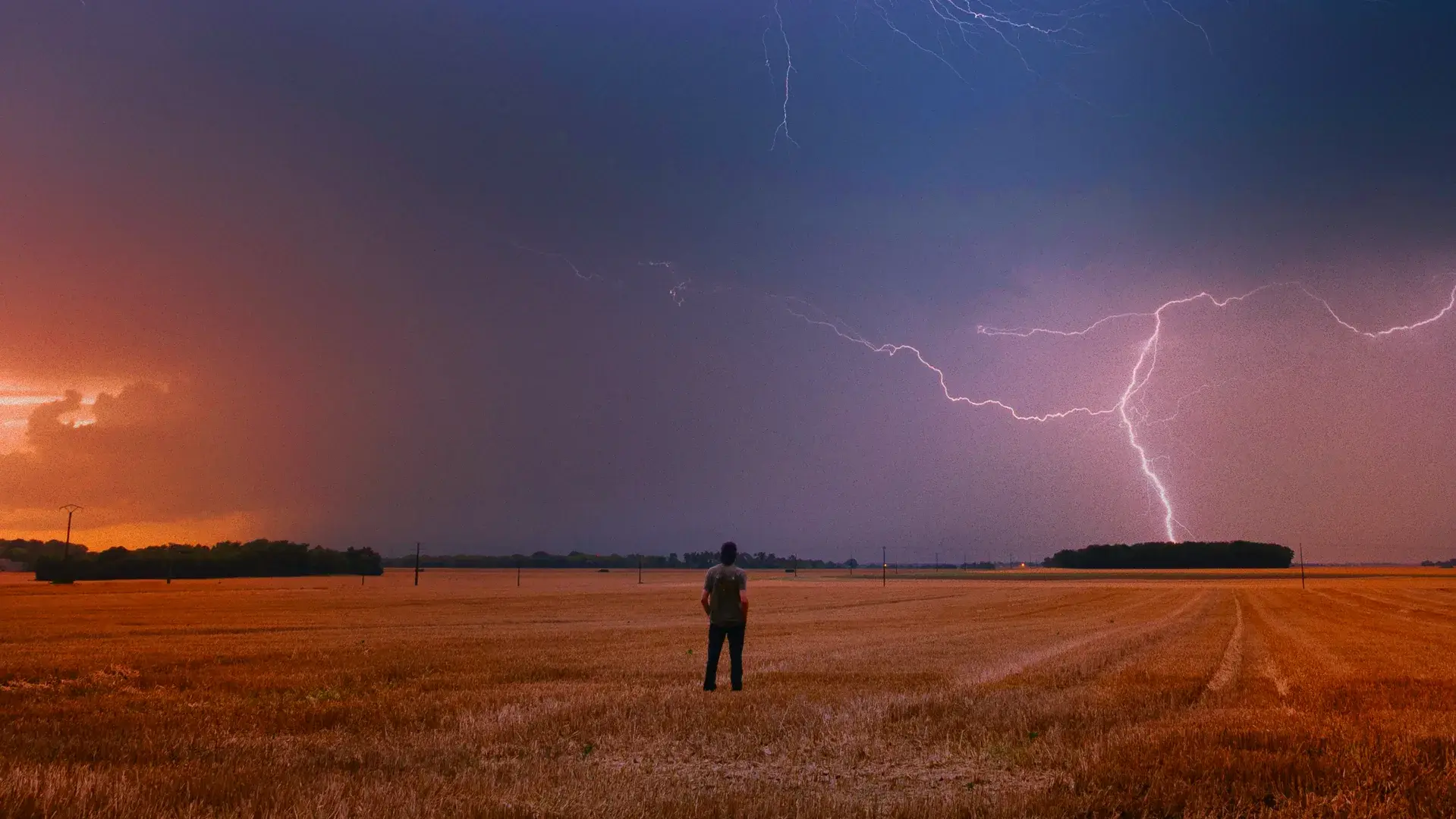 Orage au coucher du soleil au-dessus d’un champ moissonné, éclairs dans un ciel violet, un chasseur d’orages de dos au premier plan.